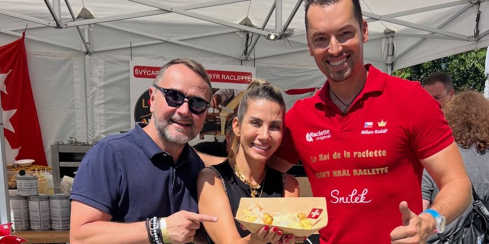 Des hôtes avec une assiette de raclette au stand Raclette Original, à côté de Milan Kolář