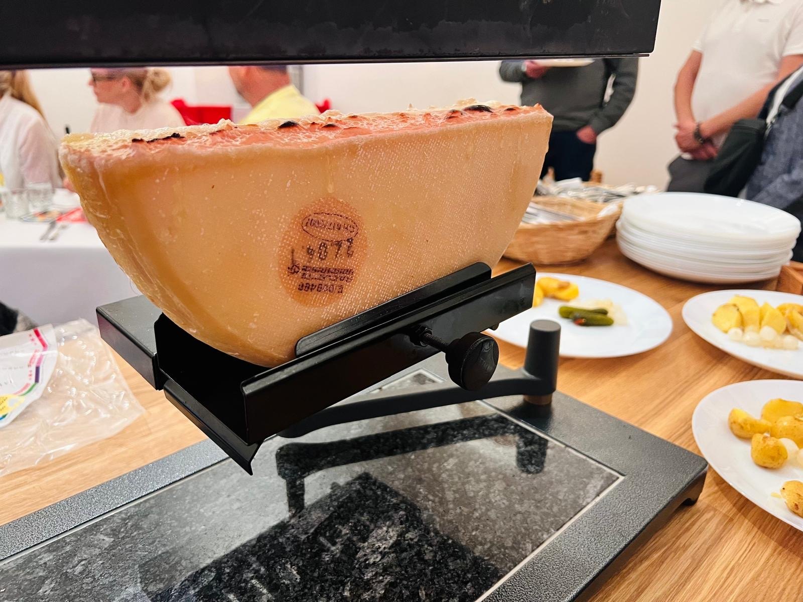 Půlka raclette sýra na grilu, vedle pečené brambory a okurky